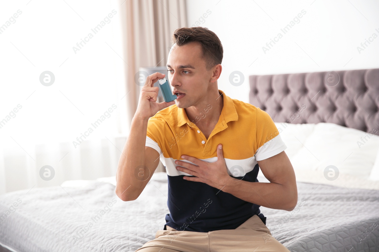 Photo of Young man with asthma inhaler on bed in light room
