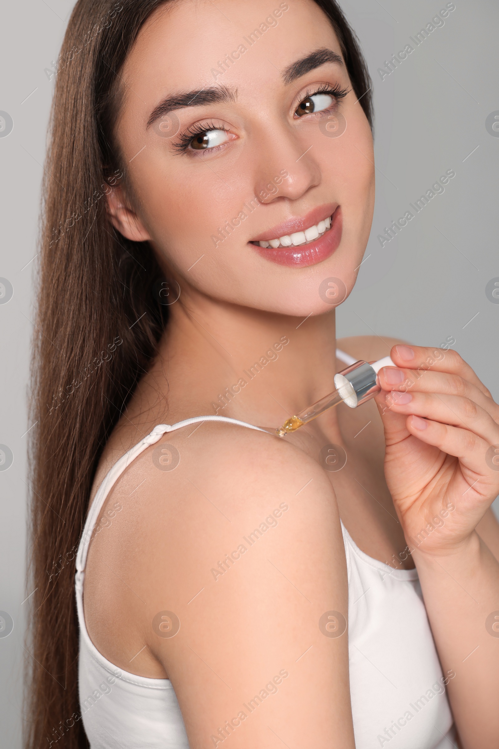 Photo of Beautiful young woman applying essential oil onto shoulder on grey background