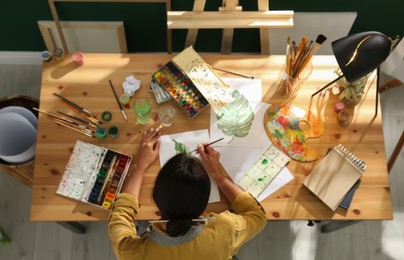 Photo of Young woman drawing leaf with watercolors at table indoors, top view