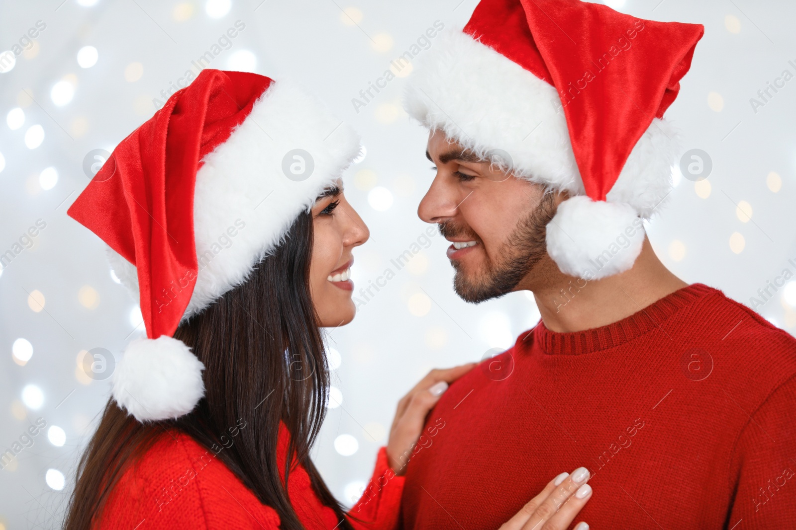 Photo of Lovely young couple in Santa hats against blurred festive lights. Christmas celebration