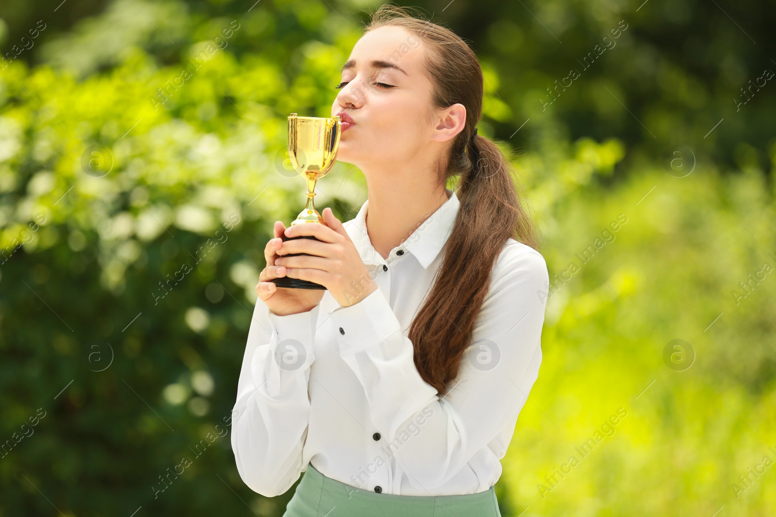 Photo of Portrait of happy young businesswoman kissing gold trophy cup in green park