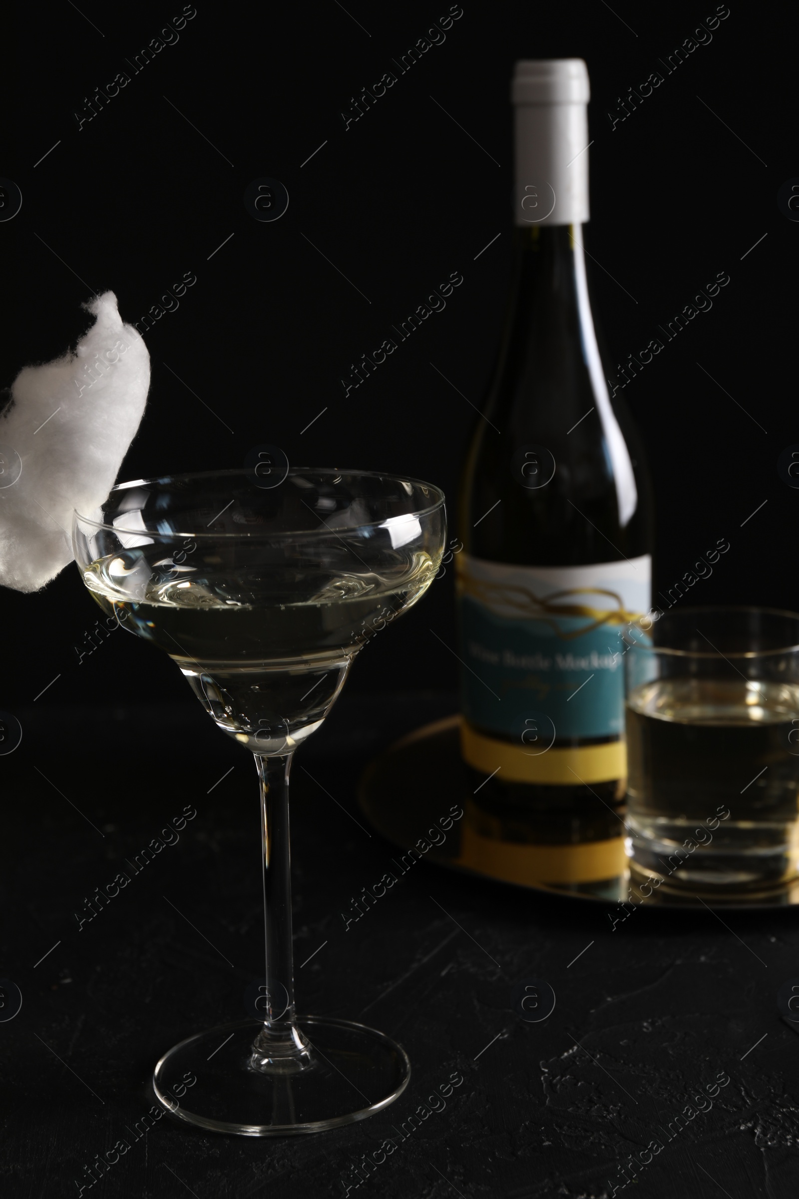 Photo of Cocktail with tasty cotton candy and bottle of alcohol drink on dark textured table against black background