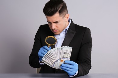 Photo of Expert authenticating 100 dollar banknotes with magnifying glass at table on light grey background. Fake money concept