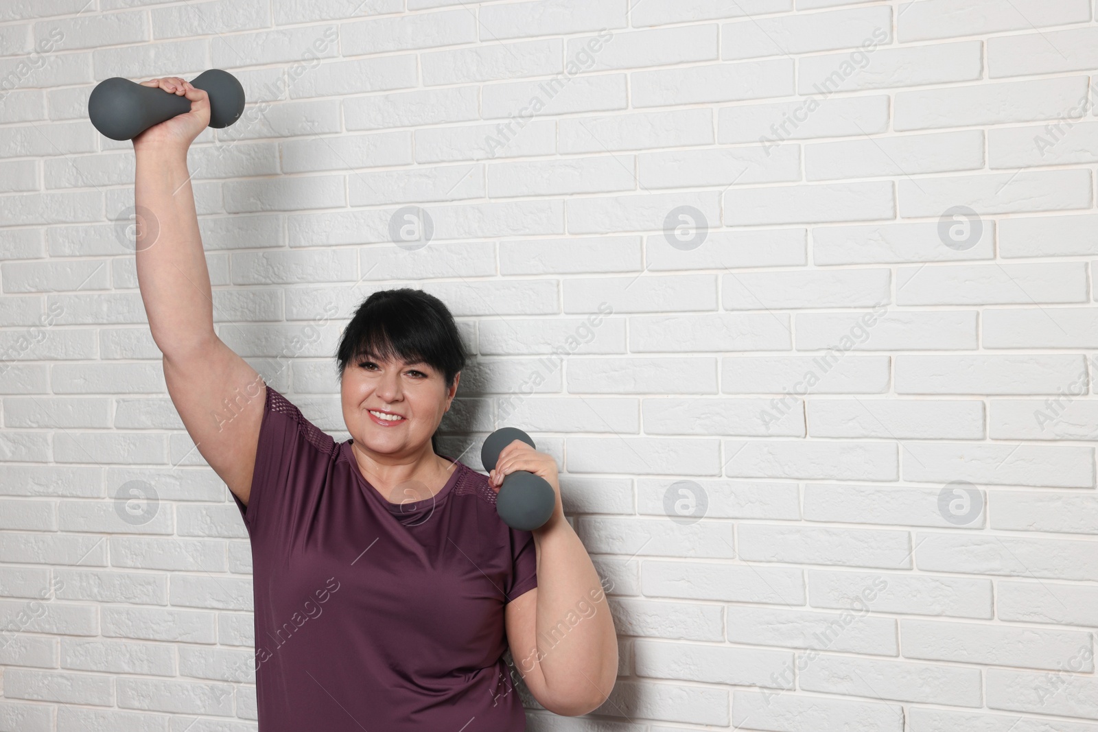 Photo of Happy overweight mature woman doing exercise with dumbbells near white brick wall, space for text
