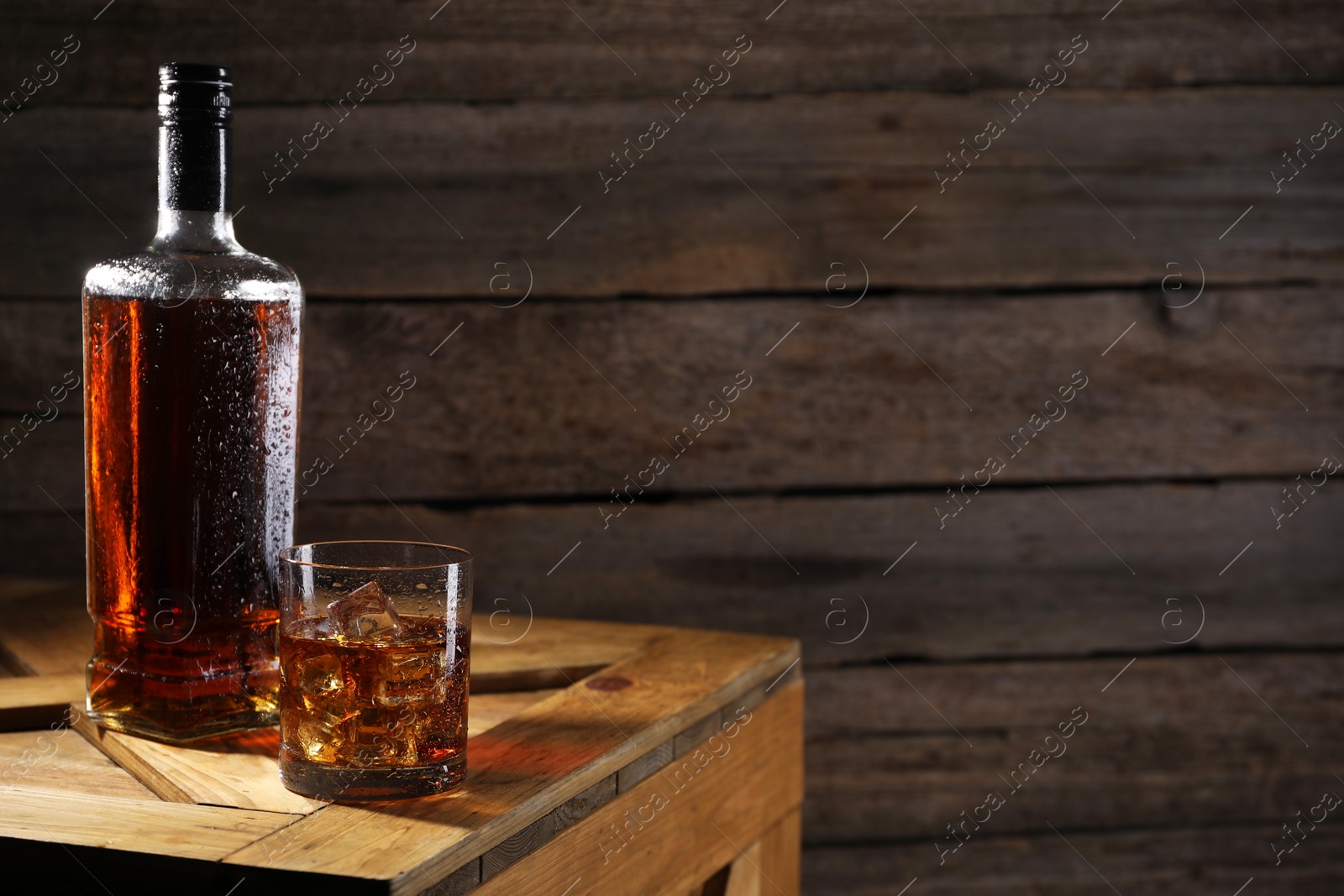 Photo of Whiskey with ice cubes in glass and bottle on wooden crate, space for text