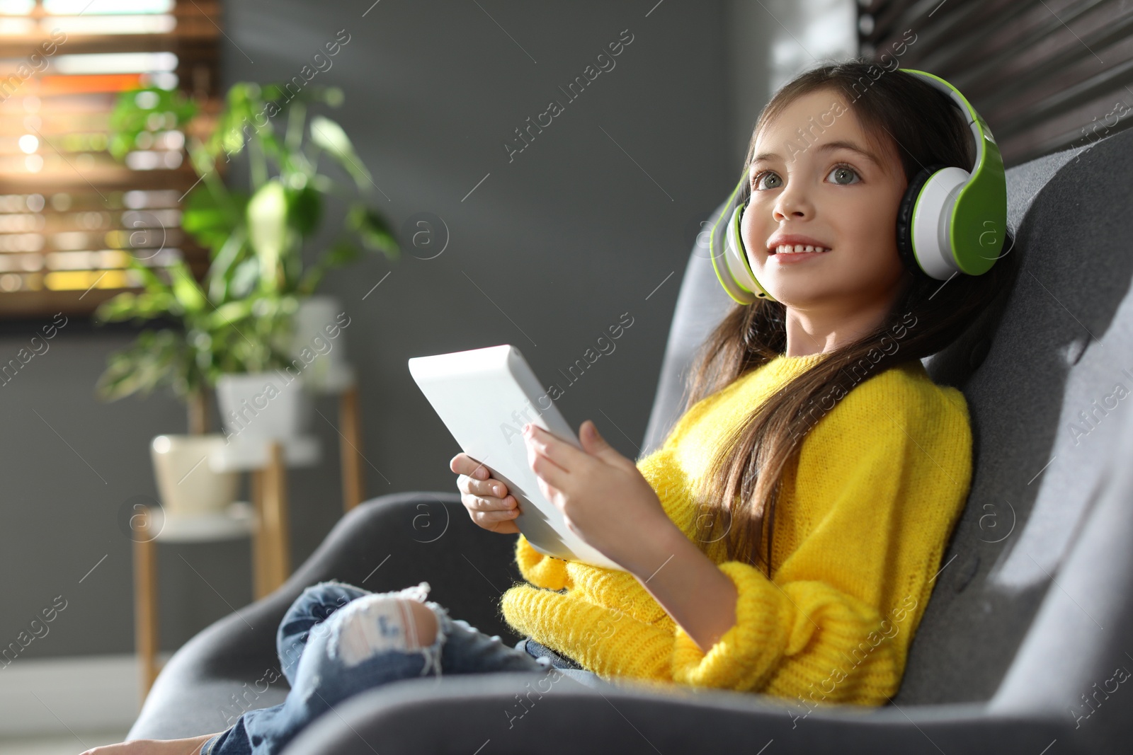 Photo of Cute little girl with headphones and tablet listening to audiobook at home