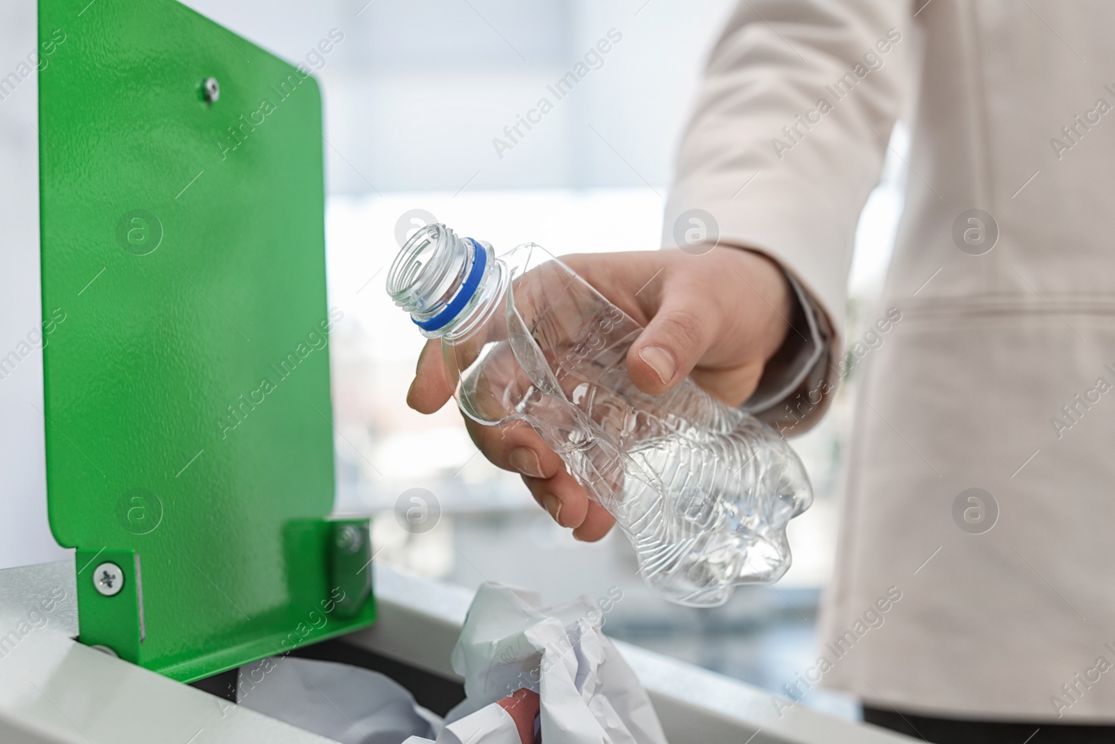 Photo of Woman putting used plastic bottle into trash bin in modern office, closeup. Waste recycling