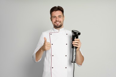 Photo of Smiling chef holding sous vide cooker and showing thumb up on beige background