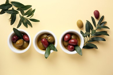 Bowls with different ripe olives and leaves on beige background, flat lay