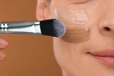 Woman applying foundation on face with brush against brown background, closeup