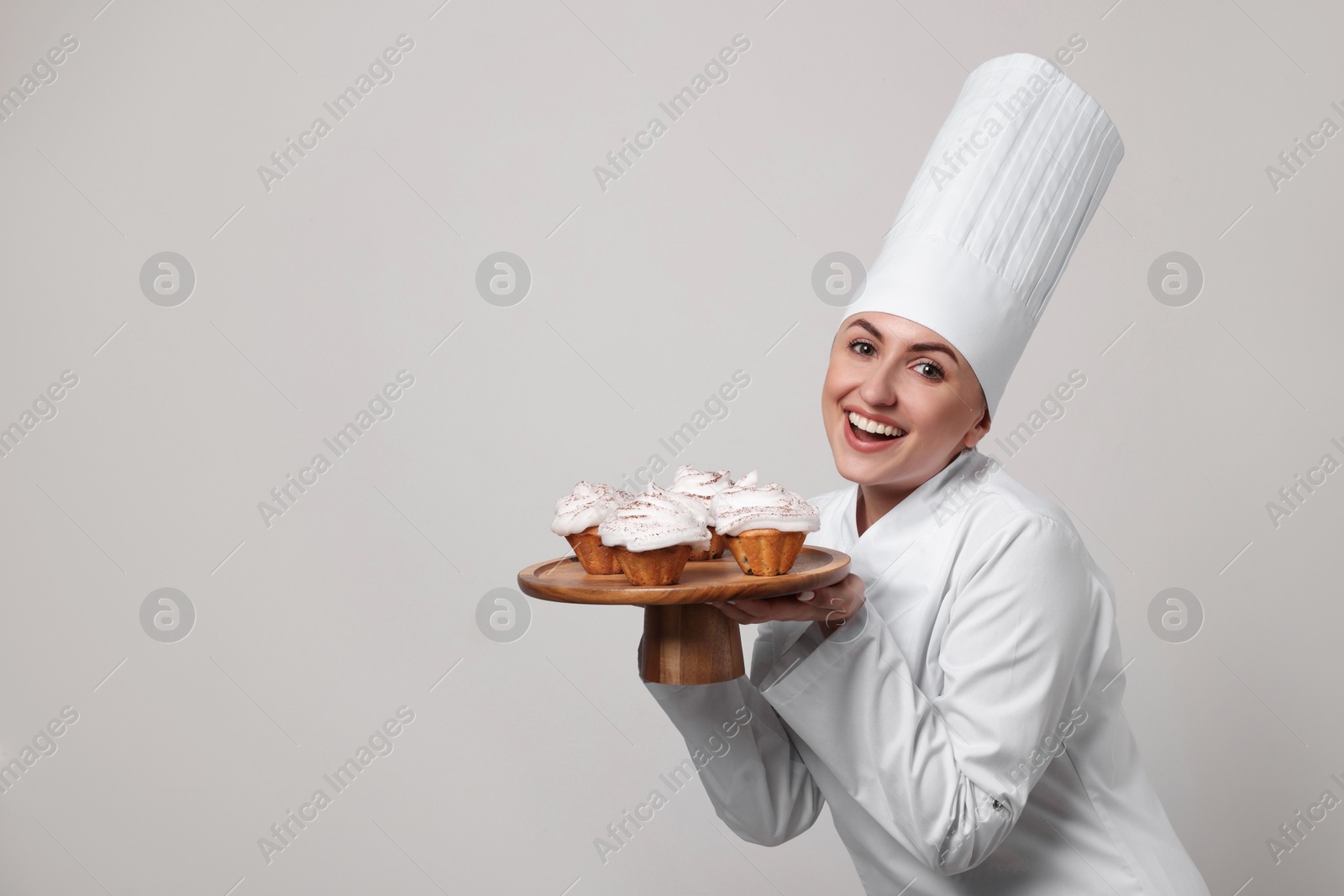 Photo of Happy professional confectioner in uniform holding delicious cupcakes on light grey background. Space for text