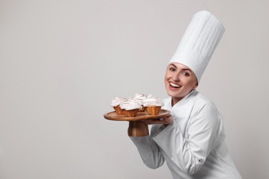 Happy professional confectioner in uniform holding delicious cupcakes on light grey background. Space for text