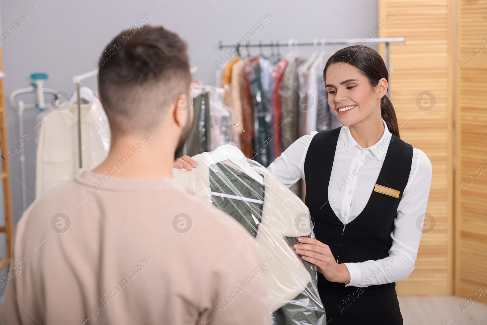 Photo of Dry-cleaning service. Happy worker giving coat in plastic bag to client indoors