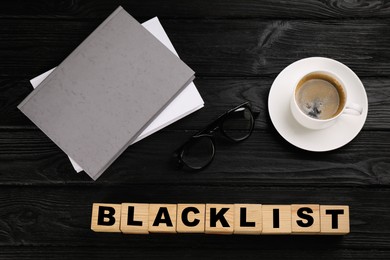 Cubes with word Blacklist, cup of coffee and office stationery on black wooden table, flat lay