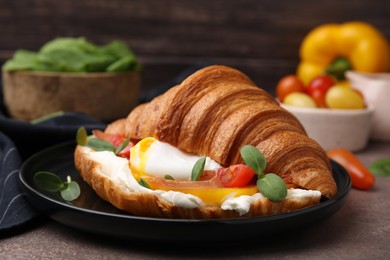 Photo of Tasty croissant with fried egg, tomato and microgreens on brown textured table, closeup