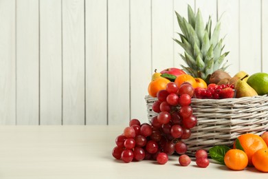 Wicker basket with different fresh fruits on white wooden table. Space for text