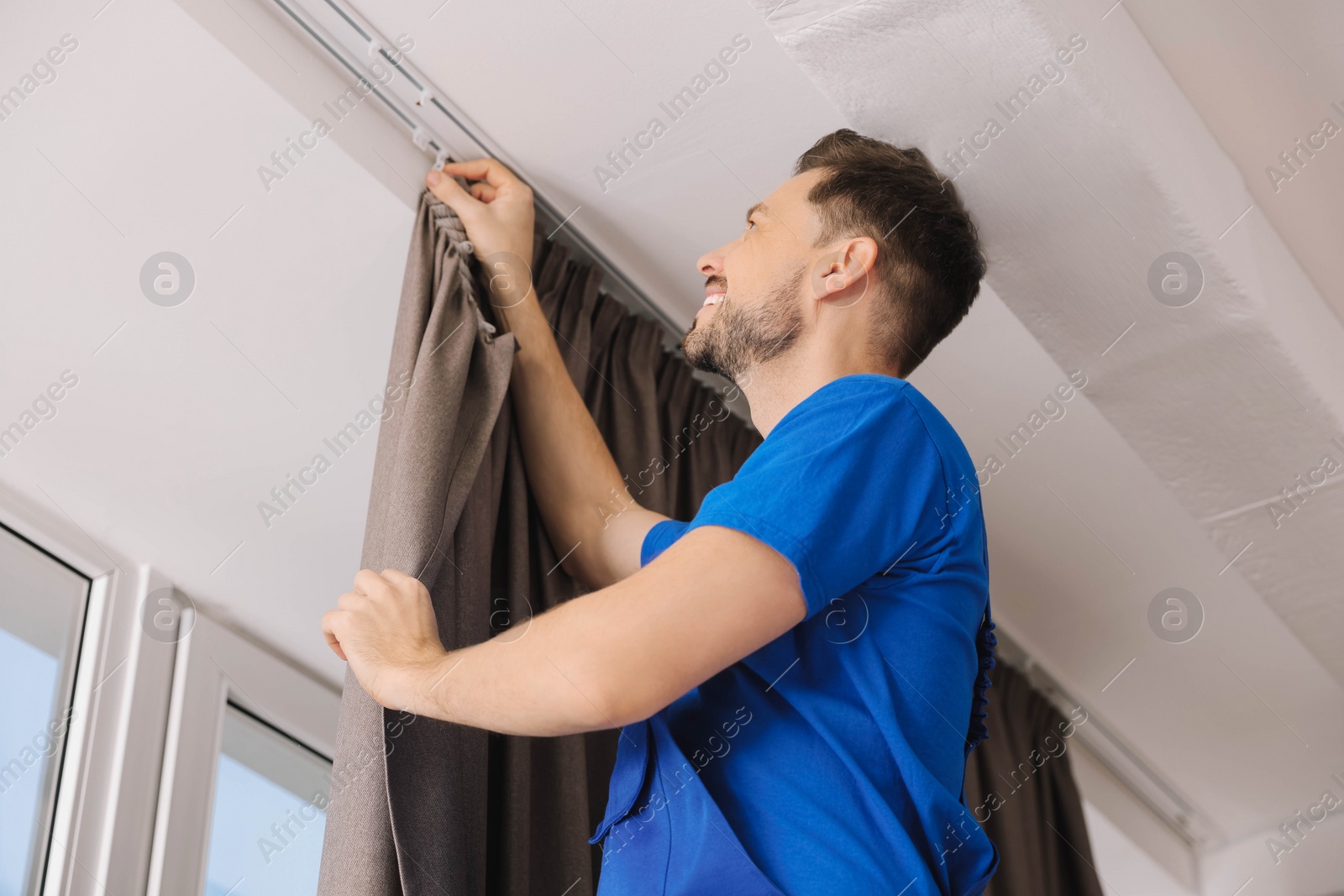 Photo of Worker in uniform hanging window curtain indoors, low angle view