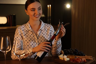 Photo of Romantic dinner. Happy woman opening wine bottle with corkscrew at table indoors