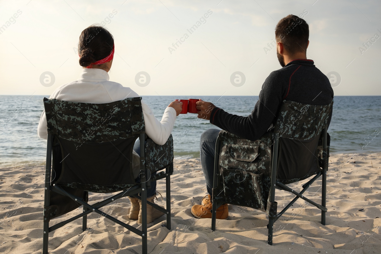 Photo of Couple sitting in camping chairs and clinking mugs on beach, back view