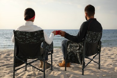 Couple sitting in camping chairs and clinking mugs on beach, back view