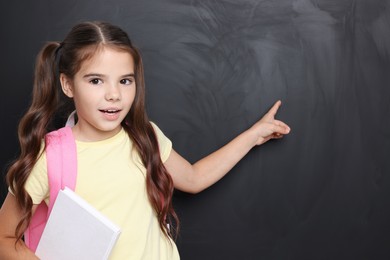 Cute schoolgirl with book near chalkboard, space for text