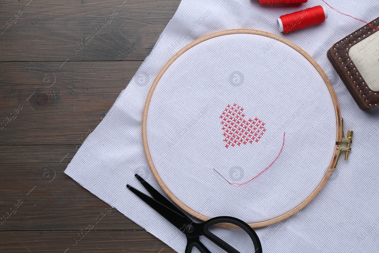 Photo of White cloth with embroidered red heart, needle and scissors on wooden table, flat lay