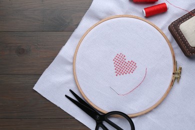 White cloth with embroidered red heart, needle and scissors on wooden table, flat lay