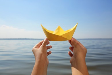 Child holding yellow paper boat near river, closeup