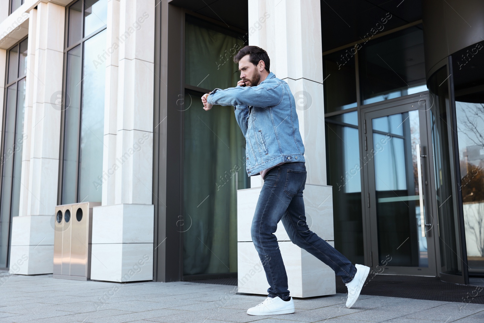 Photo of Emotional man talking on smartphone and looking at watch while running from building. Being late concept
