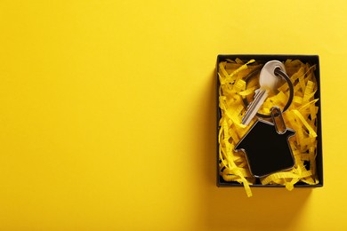 Photo of Key with trinket in shape of house and gift box on yellow background, top view with space for text. Housewarming party