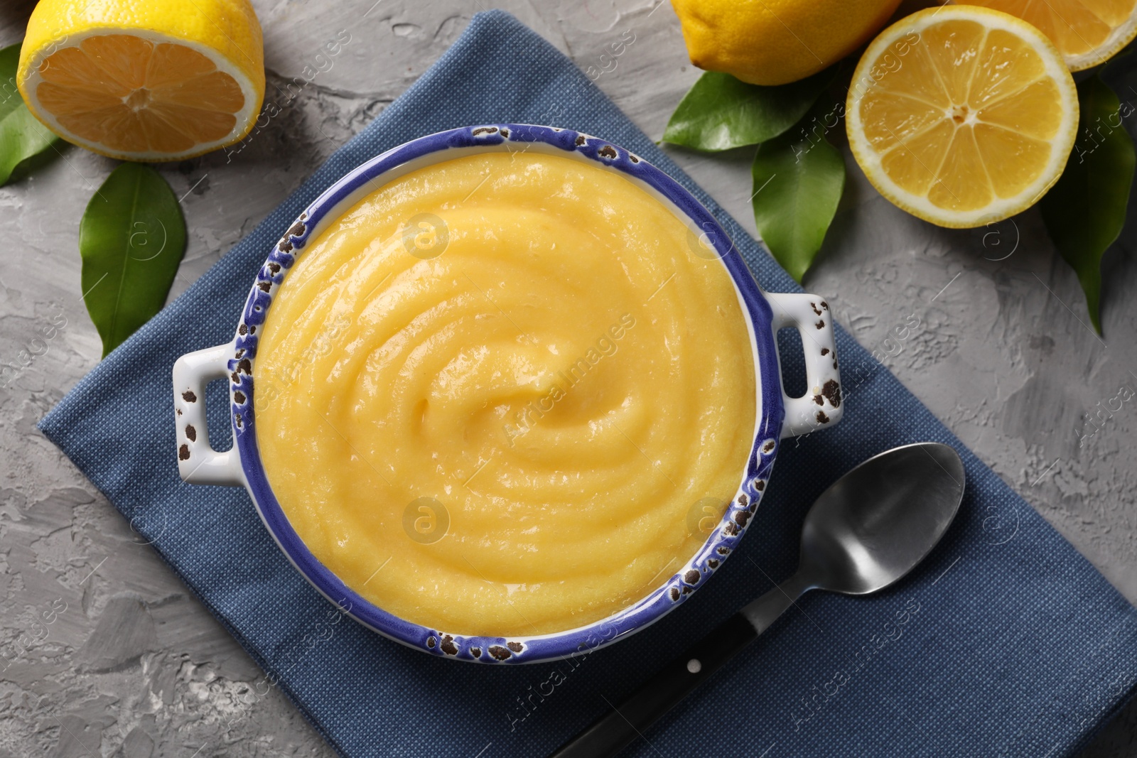 Photo of Delicious lemon curd in bowl, fresh citrus fruits, green leaves and spoon on grey table, top view