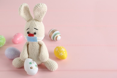 Toy bunny with protective mask and painted eggs on pink wooden table, space for text. Easter holiday during COVID-19 quarantine