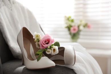 Photo of Pair of white high heel shoes, flowers and wedding dress on chair indoors