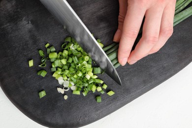 Photo of Woman cutting green spring onion on black wooden board at white table, closeup