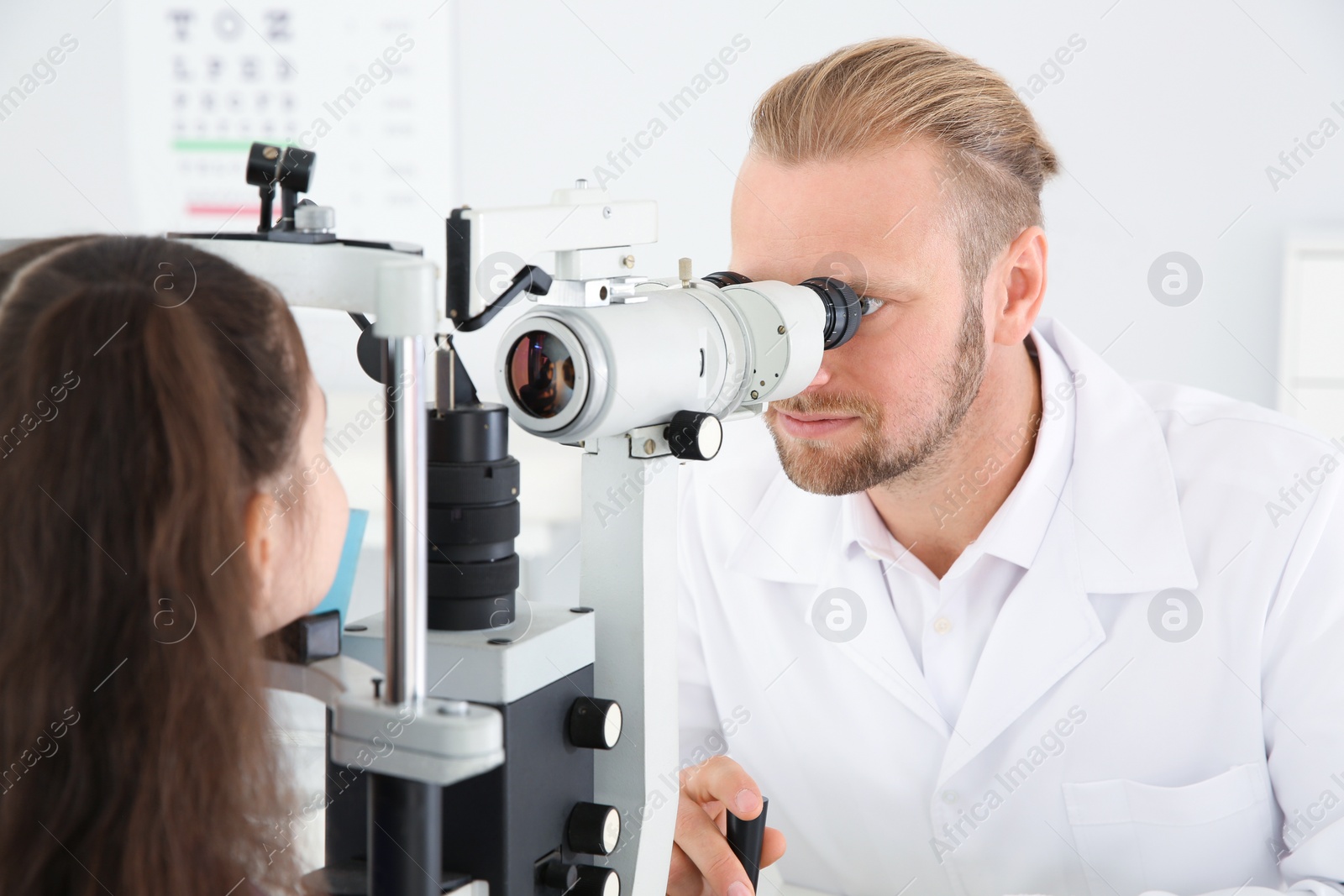 Photo of Children's doctor examining little girl with ophthalmic equipment in clinic