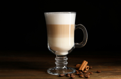 Delicious latte macchiato, cinnamon and coffee beans on wooden table against black background