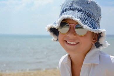 Little girl wearing sunglasses and hat at beach on sunny day. Space for text