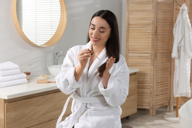 Happy young woman applying essential oil onto hair in bathroom