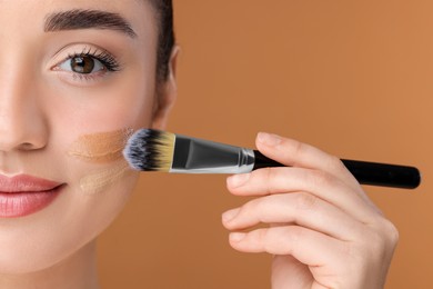 Woman applying foundation on face with brush against brown background, closeup