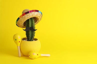 Photo of Cactus with Mexican sombrero hat, fake mustache and maracas on yellow background, space for text