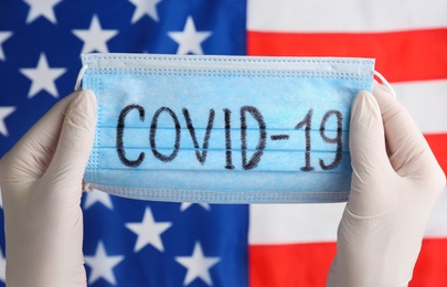 Photo of Scientist holding medical mask with word COVID-19 over American flag, closeup. Coronavirus pandemic in USA