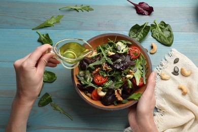 Woman adding oil to tasty fresh vegetarian salad at light blue wooden table, top view