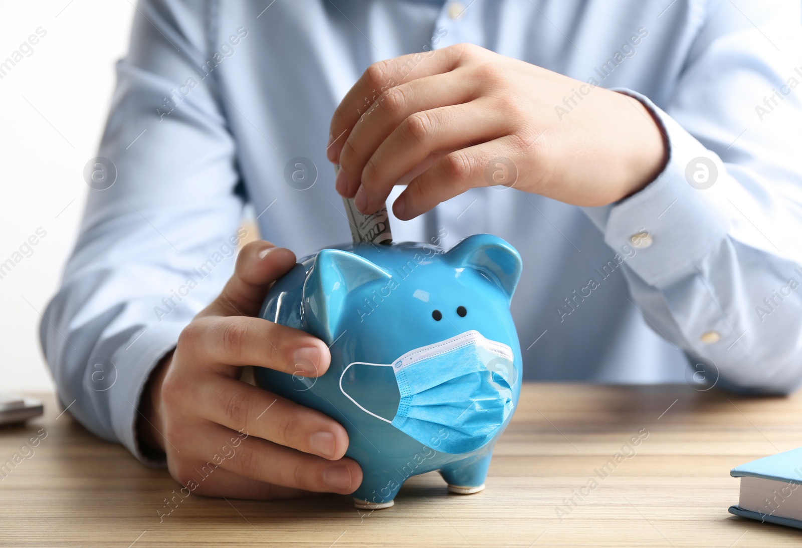 Image of Man putting dollar banknote into piggy bank with face mask at wooden table, closeup