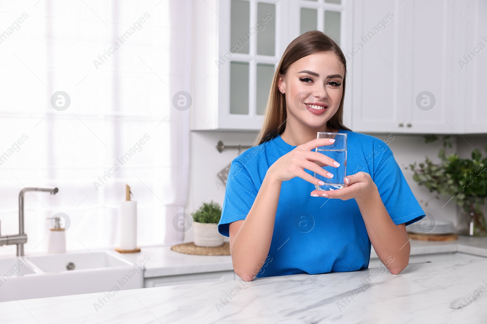 Photo of Happy woman with glass of fresh water at white marble table in kitchen, space for text