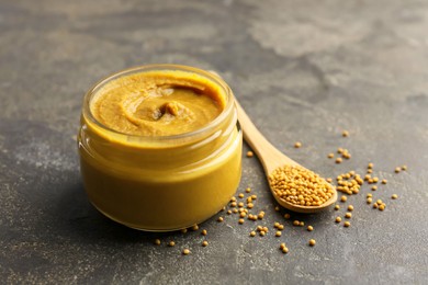 Tasty mustard sauce in jar and spoon with dry seeds on grey textured table, closeup