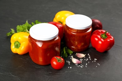 Glass jars of delicious canned lecho and fresh ingredients on black table