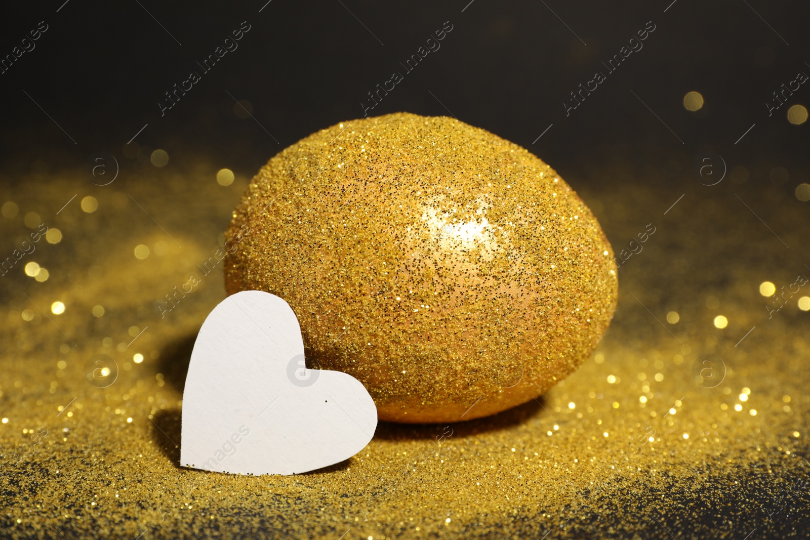 Photo of Shiny golden egg with glitter and paper heart on dark table, closeup