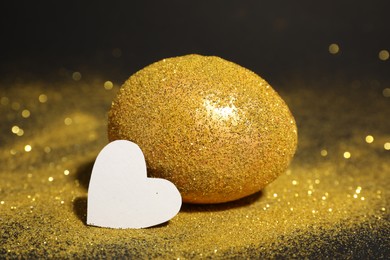 Photo of Shiny golden egg with glitter and paper heart on dark table, closeup