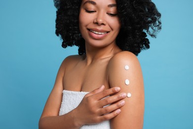 Young woman applying body cream onto shoulder on light blue background, closeup
