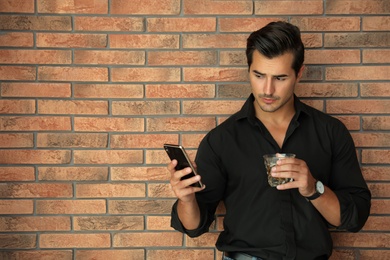 Photo of Young man with glass of whiskey and smartphone near brick wall indoors. Space for text